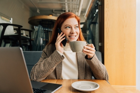 Smiling businesswoman talking on mobile phone and holding a cup of a coffee  Female in formal wear in a cafe