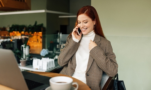 Smiling businesswoman with ginger hair talking on a smartphone  Cheerful female sitting at a table in a cafe and having a mobile phone conversation