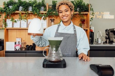 Smiling female barista holding a kettle while standing at a counter in coffee shop