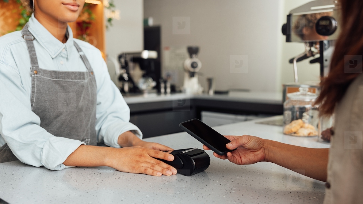 Unrecognizable customer paying by NFC at a cafe. Barista holding pos terminal while receiving payment.