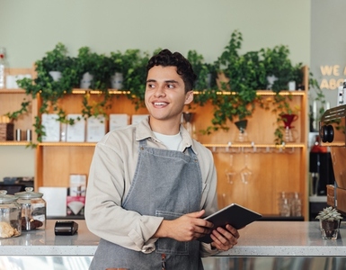 Male coffee shop owner in apron leaning counter  Middle eastern barista holding digital tablet looking away