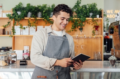 Male barista in apron holding a digital tablet while leaning on counter in coffee
