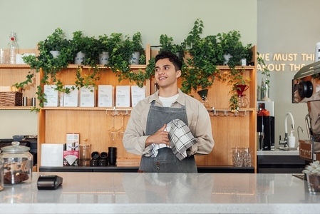 Smiling male bartender wiping a glass and looking away  Barista in a coffee shop holding a towel