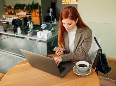 Young female entrepreneur working in a coffee shop  Woman with ginger hair typing on a laptop and holding mobile phone