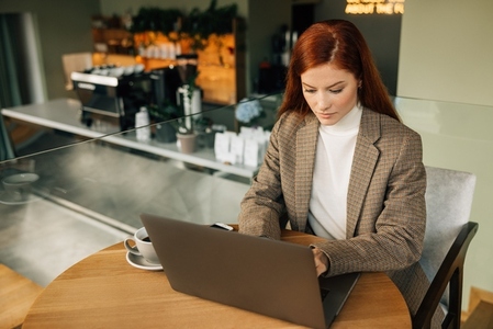 Beautiful businesswoman with ginger hair typing on a laptop while sitting at a table in a coffee shop