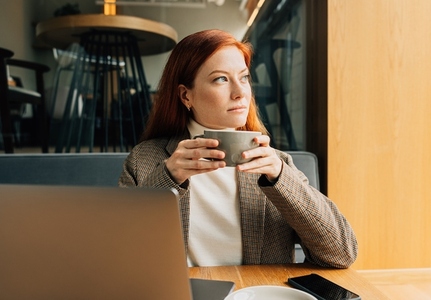 Thoughtful woman with ginger hair holding a cup of coffee sitting in a cafe looking at window