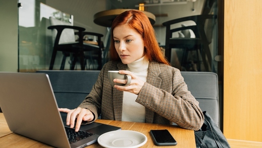Woman working as a digital nomad in a cafe  Female entrepreneur with ginger hair holding a cup typing on a laptop