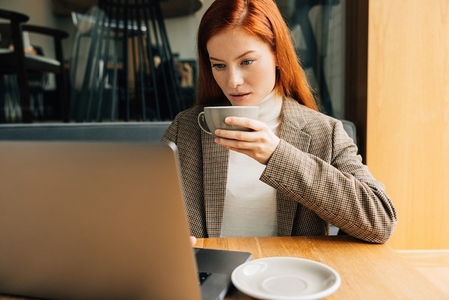 Young woman with ginger hair holding a cup of coffee and working on a laptop in cafe