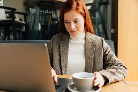 Businesswoman working in a coffee shop in the morning  Female with ginger hair in formal wear typing on laptop and drins coffee