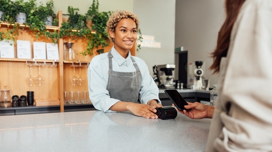 Smiling barista in apron receiving payment from a client in a coffee shop at the counter