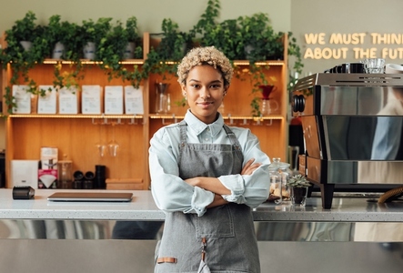Portrait of a beautiful barista in an apron  Smiling coffee shop owner with crossed arms leaning at counter