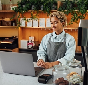 Young woman entrepreneur with laptop at the counter  Female in an apron working as a barista looking at a laptop