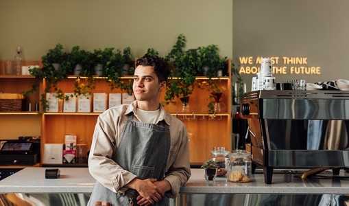 Confident local coffee shop owner leaning counter wearing an apron  Male barista looking away in a cafe