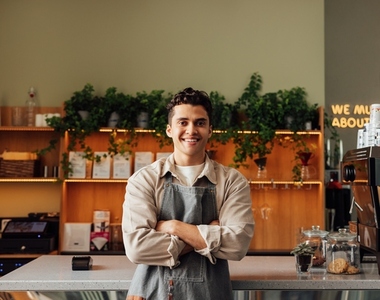 Coffee shop owner in an apron standing with crossed arms and smiling  Young handsome man leaning counter looking at a camera in a coffee shop