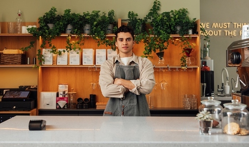 Young confident barista with cross hands looking straight at a camera while standing at a counter