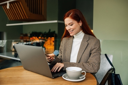 Smiling redhead woman typing on laptop and holding mobile phone while sitting in a cafe