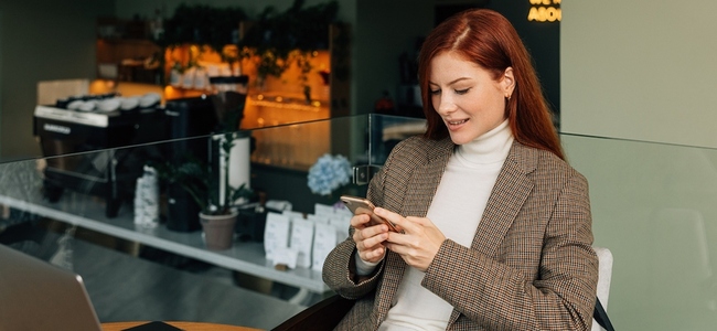 Redhead woman in formal wear sitting in a cafe holding a mobile phone and smiling