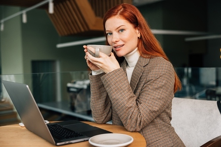 Young woman with ginger hair holding a cup  Female entrepreneur drinks coffee while working from a coffee shop