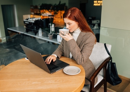 Young female entrepreneur with ginger hair typing on a laptop and drinking coffee at a cafe