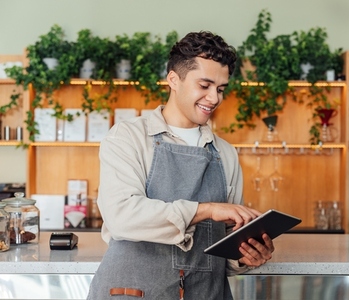 Smiling barista in an apron holding a digital tablet while leaning counter  Coffee shop owner typing on digital tablet