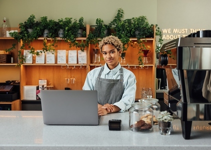 Young female entrepreneur in an apron looking at camera while standing at the counter with laptop and coffee machine