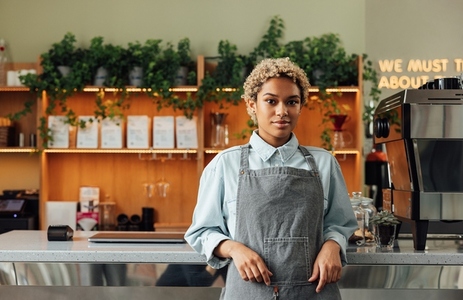 Confident female barista leaning on a counter looking at camera  Young woman in an apron working as a barista standing in a cafe