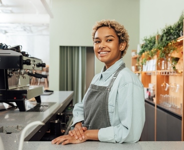 Young smiling barista in an apron leaning counter in a coffee shop  Portrait of a cheerful female bartender