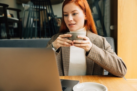 Beautiful woman with ginger hair holding a cup of coffee with two hands looking at a laptop
