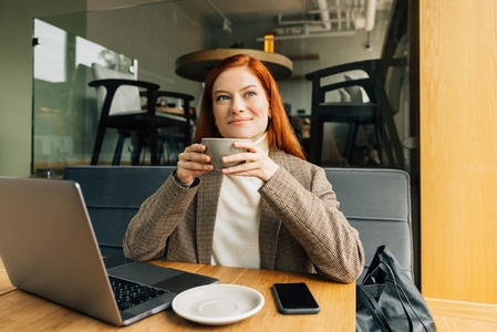 Smiling redhead woman in formal wear drinking coffee looking at window in cafe