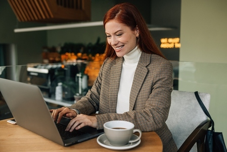 Cheerful woman with ginger hair working in a cafe  Smiling female in formal wear sitting at a table in a coffee shop