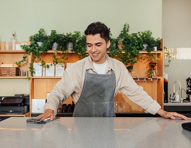 Smiling barista cleaning a counter in a cafe with a towel  Young male in an apron working as a bartender