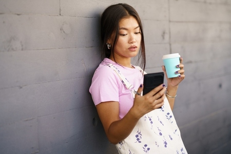 Asian woman with coffee using smartphone near wall
