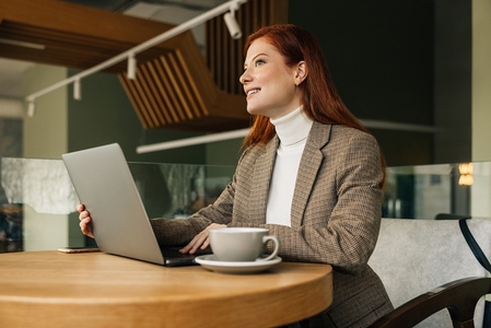 Woman entrepreneur sitting at a table with a laptop  Redhead female working remotely from a cafe