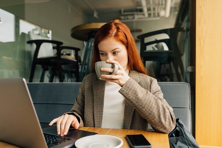 Young female with ginger hair drinking from a cup and typing on a laptop