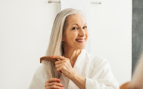 Cheerful aged female combing her gray hair in front of a bathroom mirror
