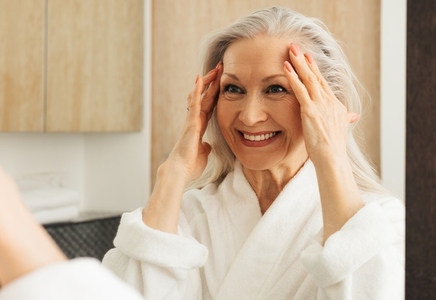 Happy senior woman with grey hair looking at a mirror and touching her face while looking at mirror