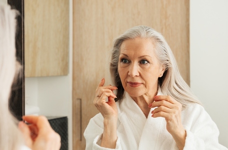 Senior woman examines her face in front of bathroom mirror
