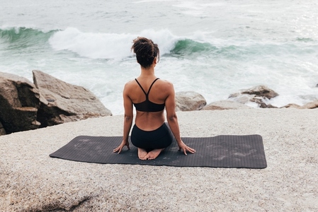 Back view of a fit woman in sports clothes sitting on a mat and looking at waves during a yoga session