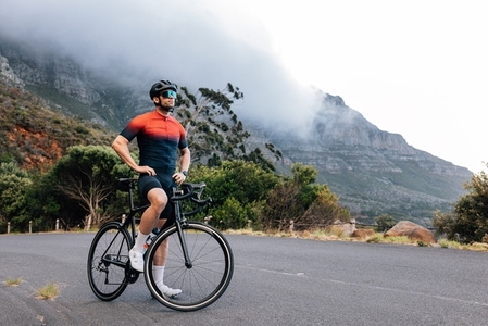 Young male cyclist relaxing after training with hands on a hips  Professional athlete with road bike enjoying the view while standing on a road in wild terrain