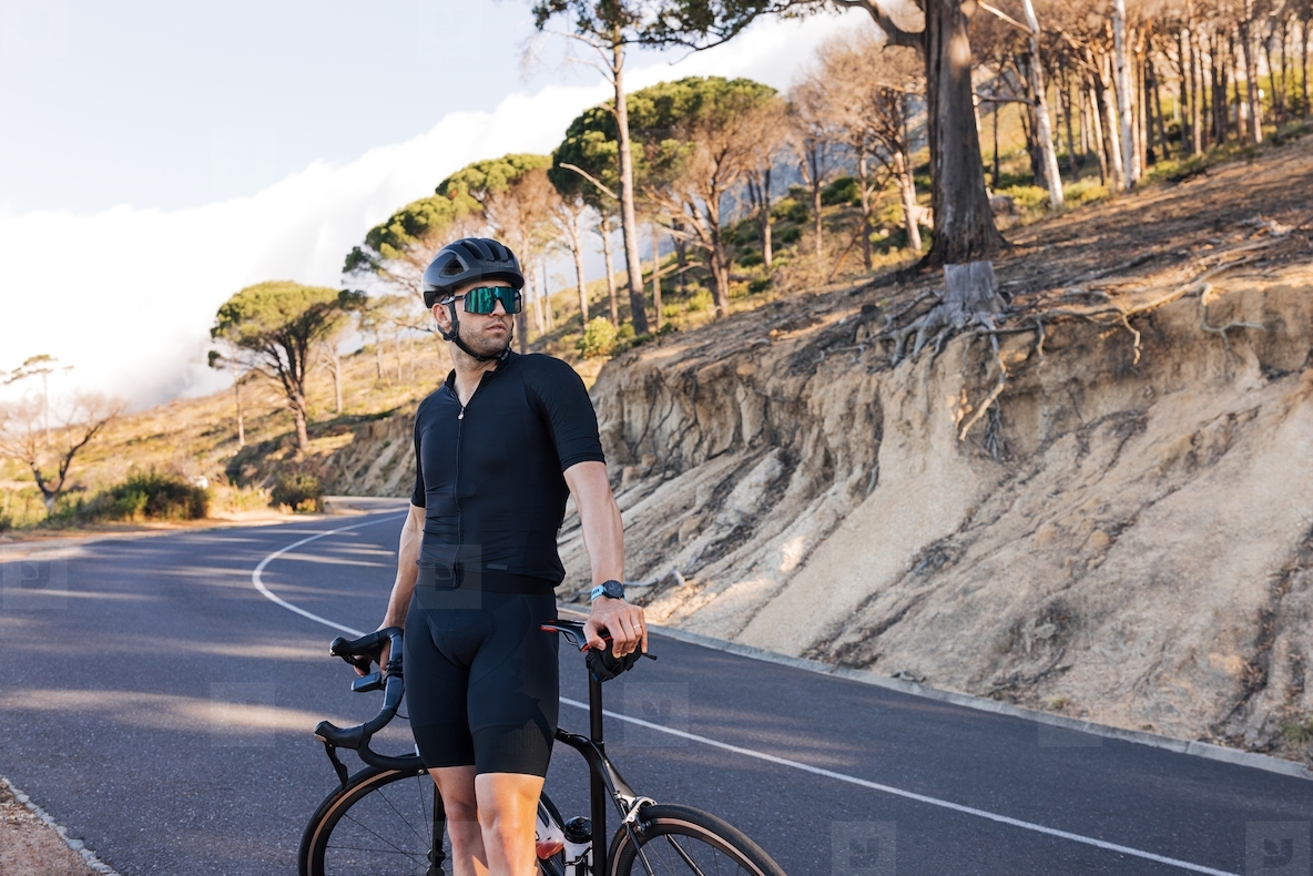Portrait of a professional cyclist in a helmet looking away while standing on a road. Athlete in black sportswear with road bike relaxing on a country road