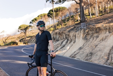 Portrait of a professional cyclist in a helmet looking away while standing on a road  Athlete in black sportswear with road bike relaxing on a country road