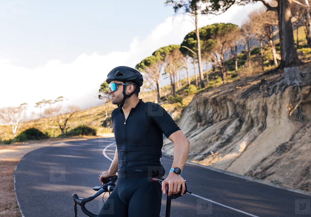 Side view of a professional biker relaxing leaning on his road bike. Male cyclist in glasses and helmet enjoying the view and taking a break during the ride