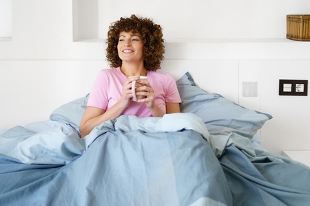 Smiling woman with cup of hot drink on bed