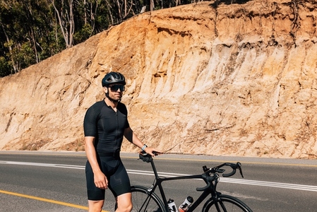 Young male cyclist in black sportswear walking on a road with his bicycle