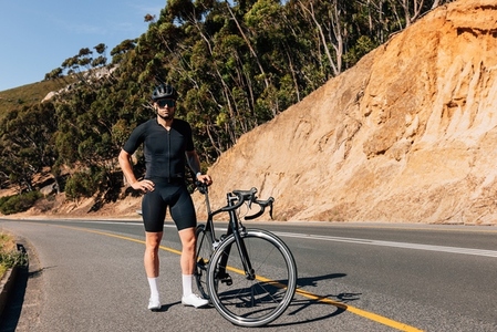 Professional cyclist in black sportswear with his road bike looking at a camera while standing on an empty road