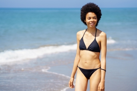 Content black woman in bikini walking on seashore