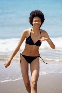 Joyful black woman in bikini walking along seaside