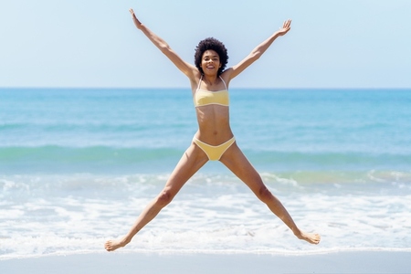 Excited ethnic woman jumping with raised arms on sandy seashore