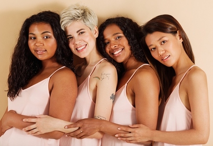 Group of smiling females with different skin tones against a beige background  Different women standing together in studio  Diverse cheerful females are hugging each other