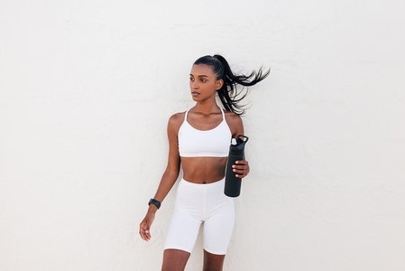 Portrait of a slim sportswoman with a shaker at a white wall  Female in white fitness attire holding a bottle